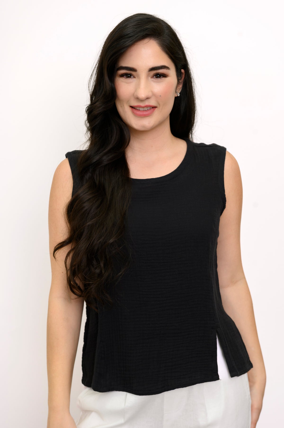 Woman wearing a black sleeveless top and white pants against a white background, front view