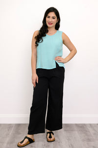 Woman wearing a light blue sleeveless top, black pants and black sandals  against a white wall.