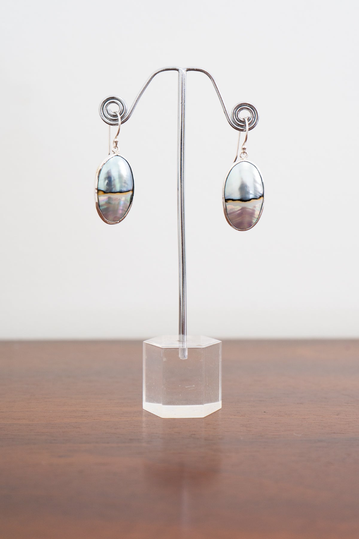 Pair of silver pearl shell earrings on a clear stand with a wooden surface and white background, front view
