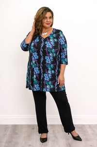 Plus-size model wearing a blue-teal floral A-line tunic and black fitted pants, standing against a white wall.