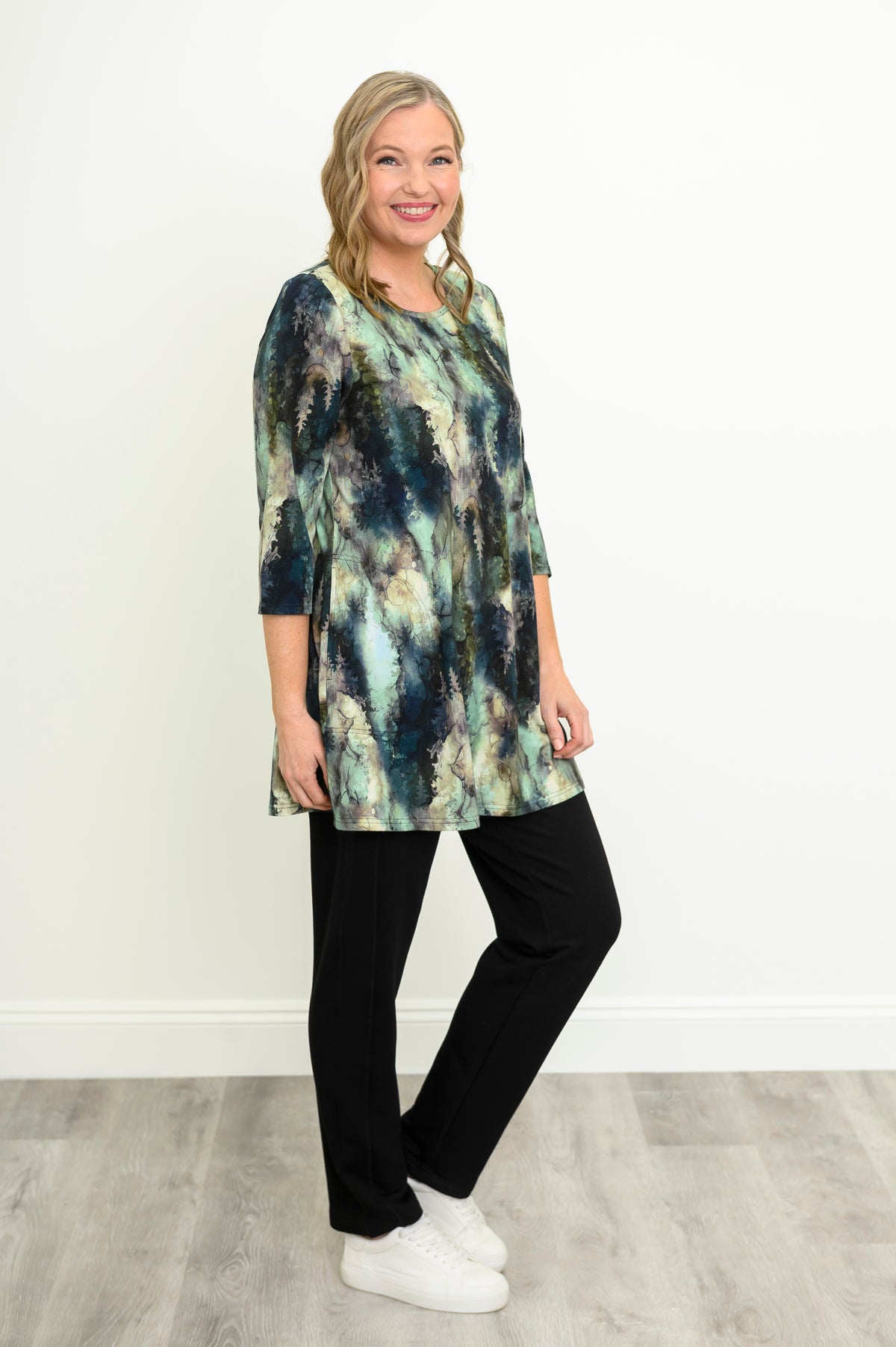 Woman in a blue-green watercolor forest print A-line tunic, black trousers, and white sneakers against a white wall.