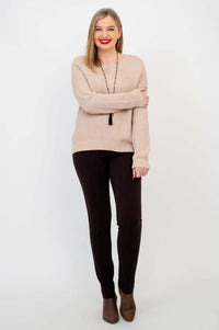 Woman in beige knit sweater and brown pants posing with arm across body