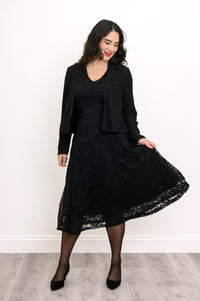 Female model playing with the hem of her black lace flared dress, smiling, against white wall and gray floor