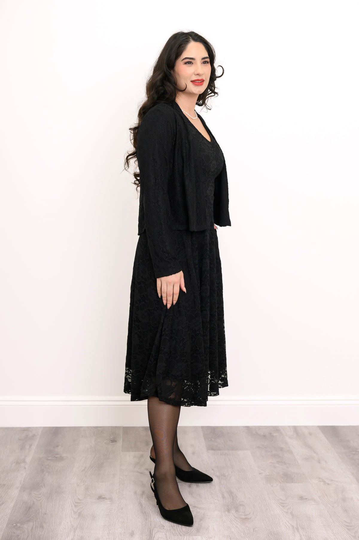 Side view of female model wearing loose-fit black dress with lace overlay and matching cardigan, against white wall and gray floor