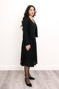 Side view of female model wearing loose-fit black dress with lace overlay and matching cardigan, against white wall and gray floor