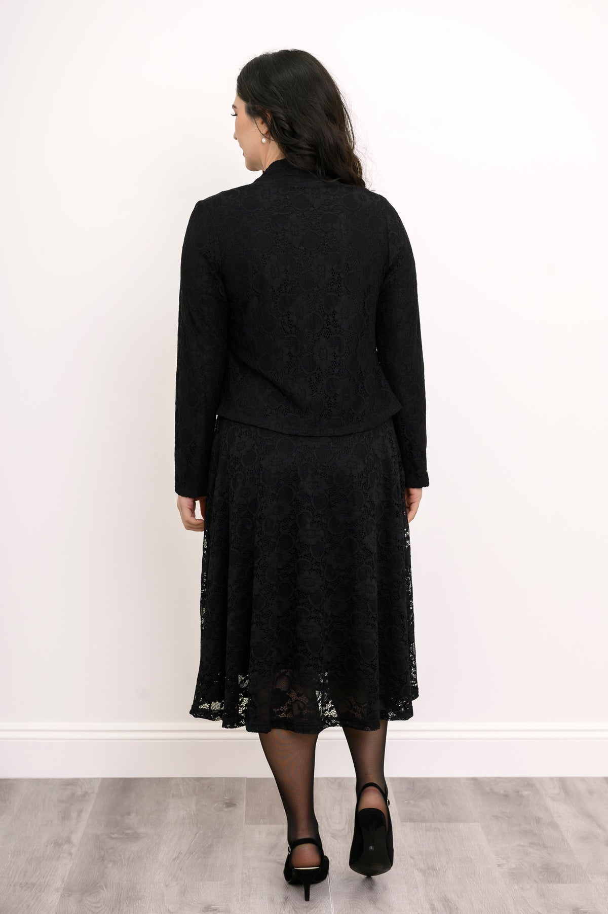 Back view of female model standing in formal black lace outfit featuring a matching cardigan and dress, against white wall and gray floor