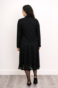 Back view of female model standing in formal black lace outfit featuring a matching cardigan and dress, against white wall and gray floor