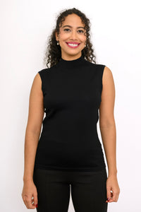 A woman with curly dark hair wearing a black sleeveless mock neck top and black pants, standing against a plain white studio background.