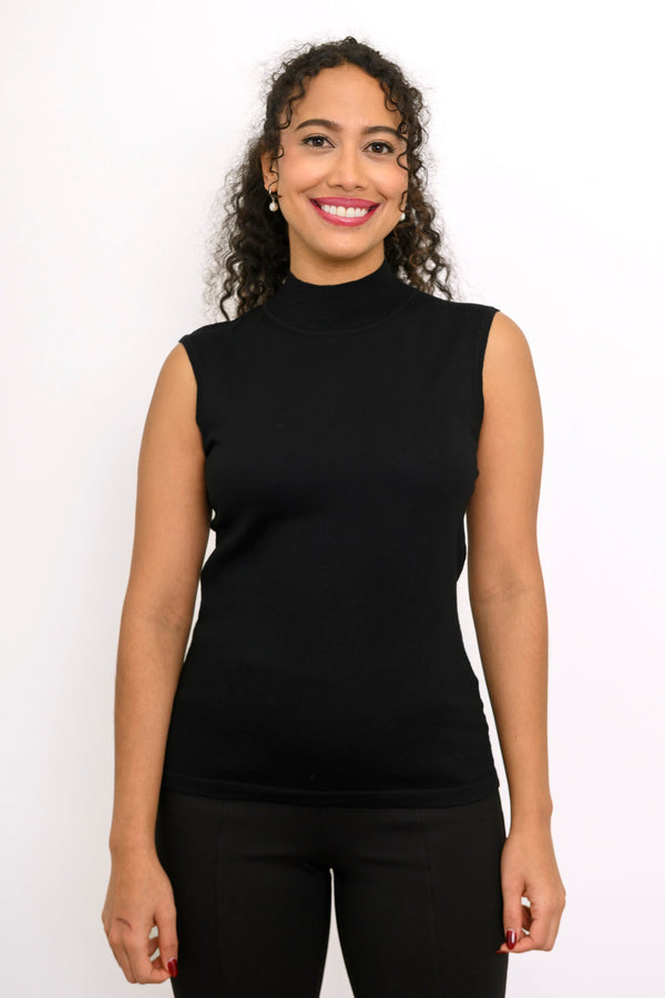 A woman with curly dark hair wearing a black sleeveless mock neck top and black pants, standing against a plain white studio background.