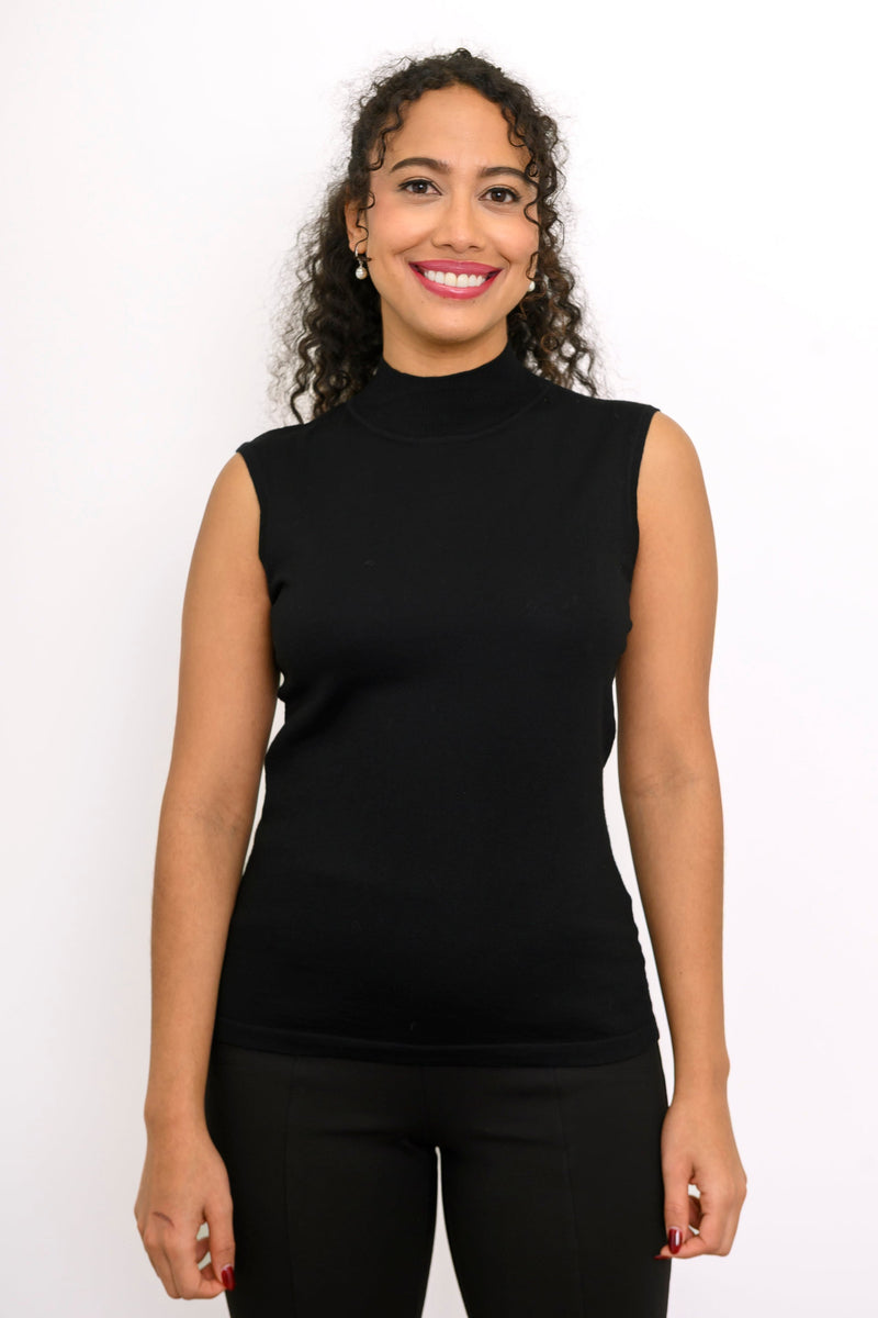 A woman with curly dark hair wearing a black sleeveless mock neck top and black pants, standing against a plain white studio background.