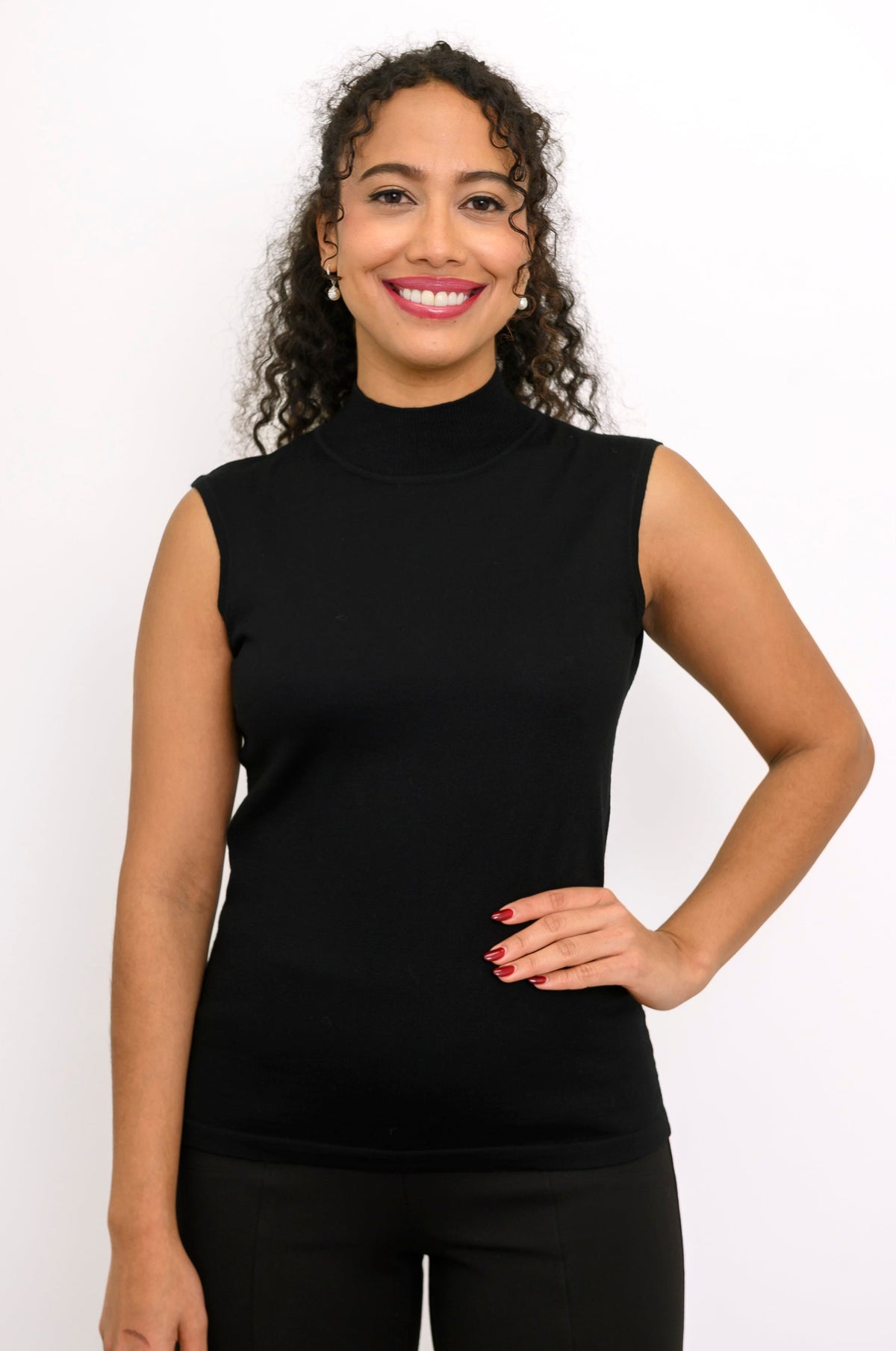 A smiling woman with curly hair wearing a black sleeveless mock neck top and black pants, posing against a plain white background.