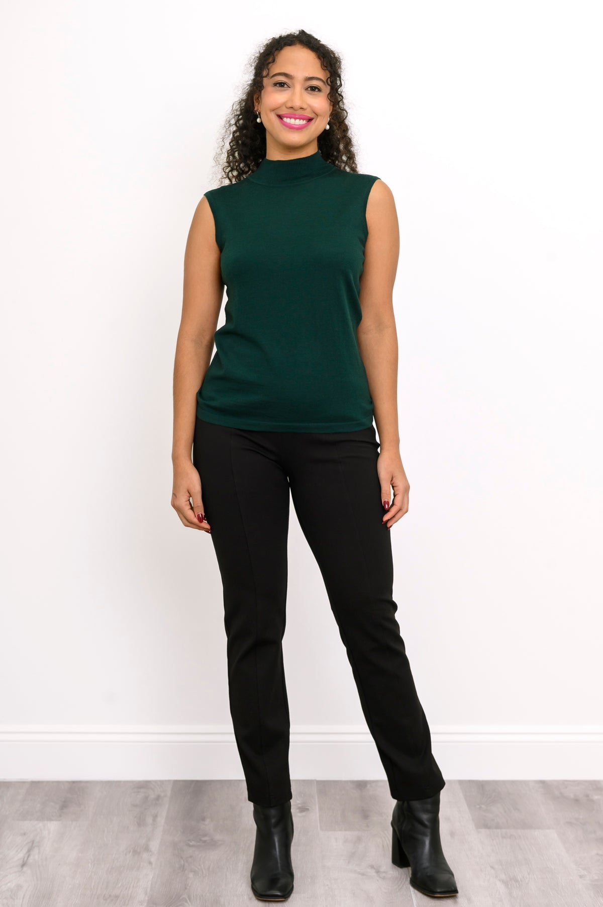 Full length view of a woman wearing a deep forest green sleeveless merino wool knit top and black pants.