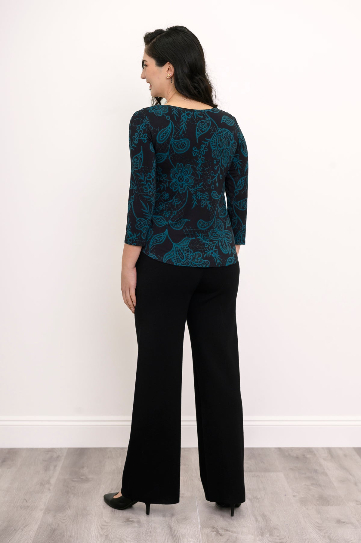 Back view of a women's A-line teal leaf print top styled with dark trousers against a white wall.