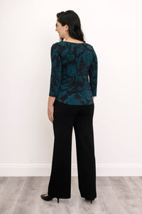 Back view of a women's A-line teal leaf print top styled with dark trousers against a white wall.