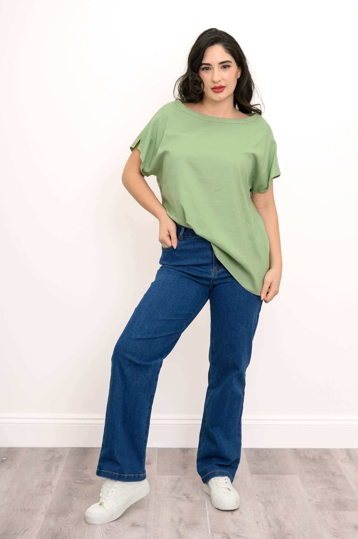 Woman posing in a sage green boatneck tee, blue jeans, and white sneakers on a light wood floor
