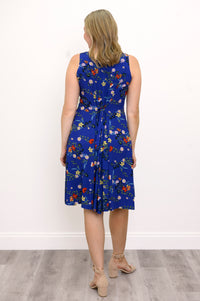 Back view of woman wearing a blue floral dress and sandals against a white wall 