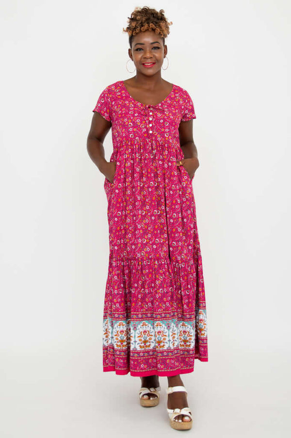 Woman showing pockets in a pink floral maxi dress with a tiered skirt and white sandals