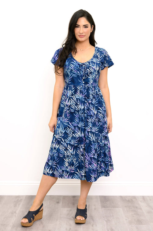A woman modeling a blue midi dress with a white tropical leaf print, standing against a white wall on a light gray wood floor.