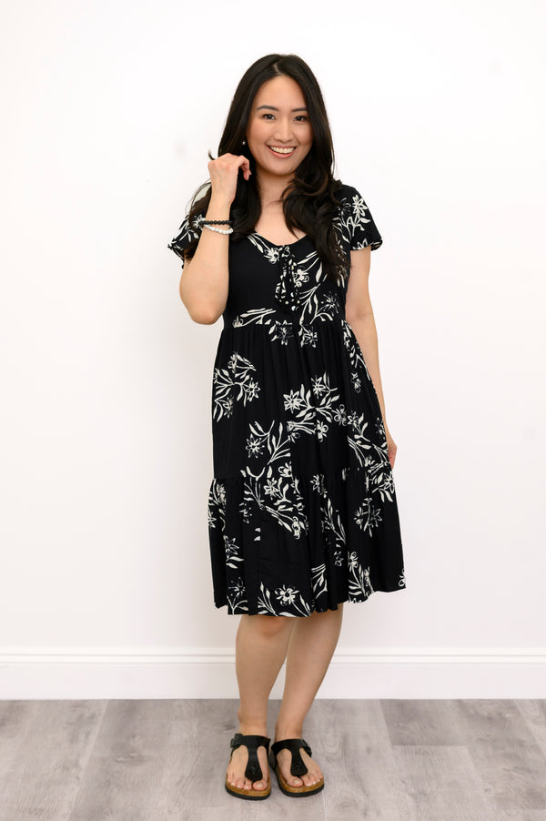Full body view of woman with her hand near her face and smiling while wearing a floral dress and sandals
