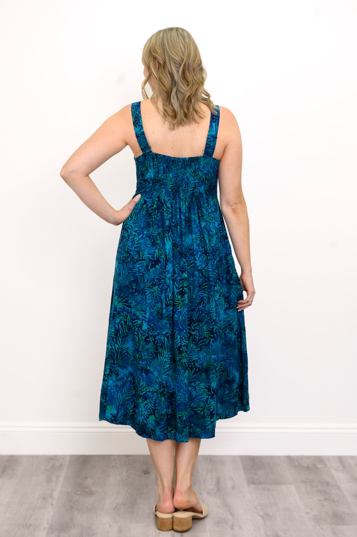 Woman wearing a blue patterned dress with a ruched back and sandals against a white wall, back view