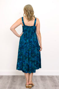 Woman wearing a blue patterned dress with a ruched back and sandals against a white wall, back view