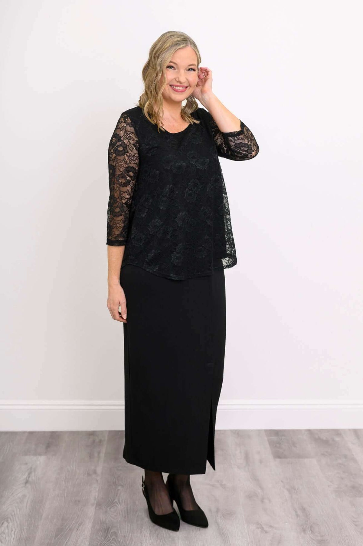 Woman modeling black lace overlay top paired with a black skirt, touching her hair