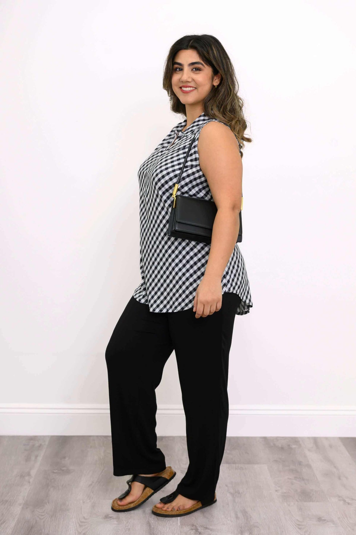 Side view of a woman in a gingham tank top, black pants, sandals, and shoulder bag