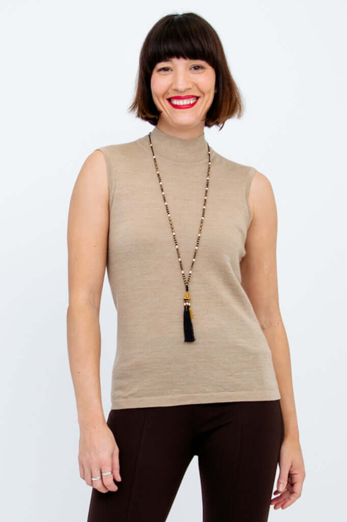 Woman wearing a sleeveless beige mock neck sweater and brown pants, smiling