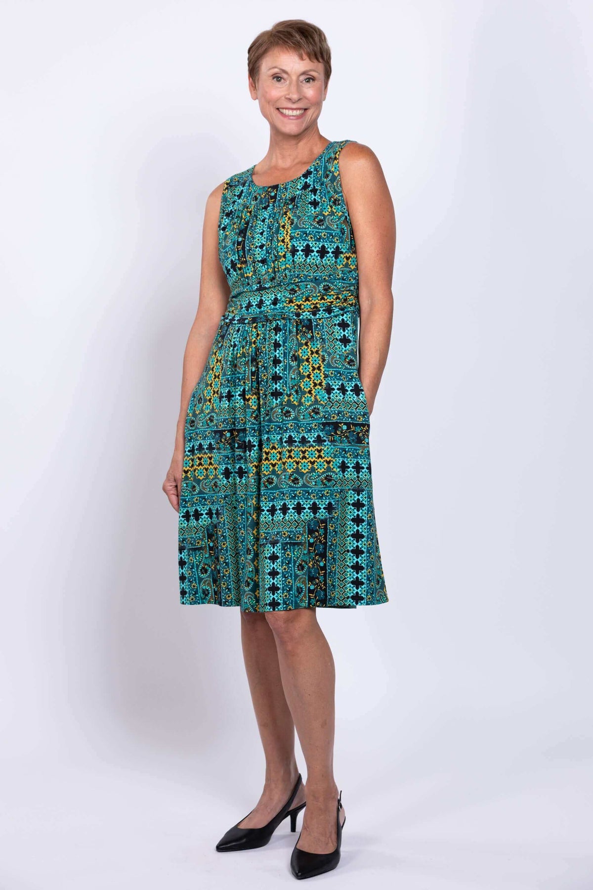 Woman in sleeveless turquoise mosaic dress with hands in pockets, side angle