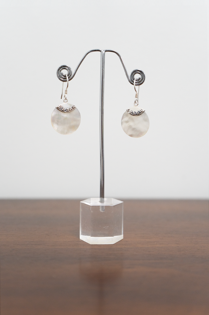 A pair of sterling silver earrings with a flat round pearl pendant displayed on a metal stand