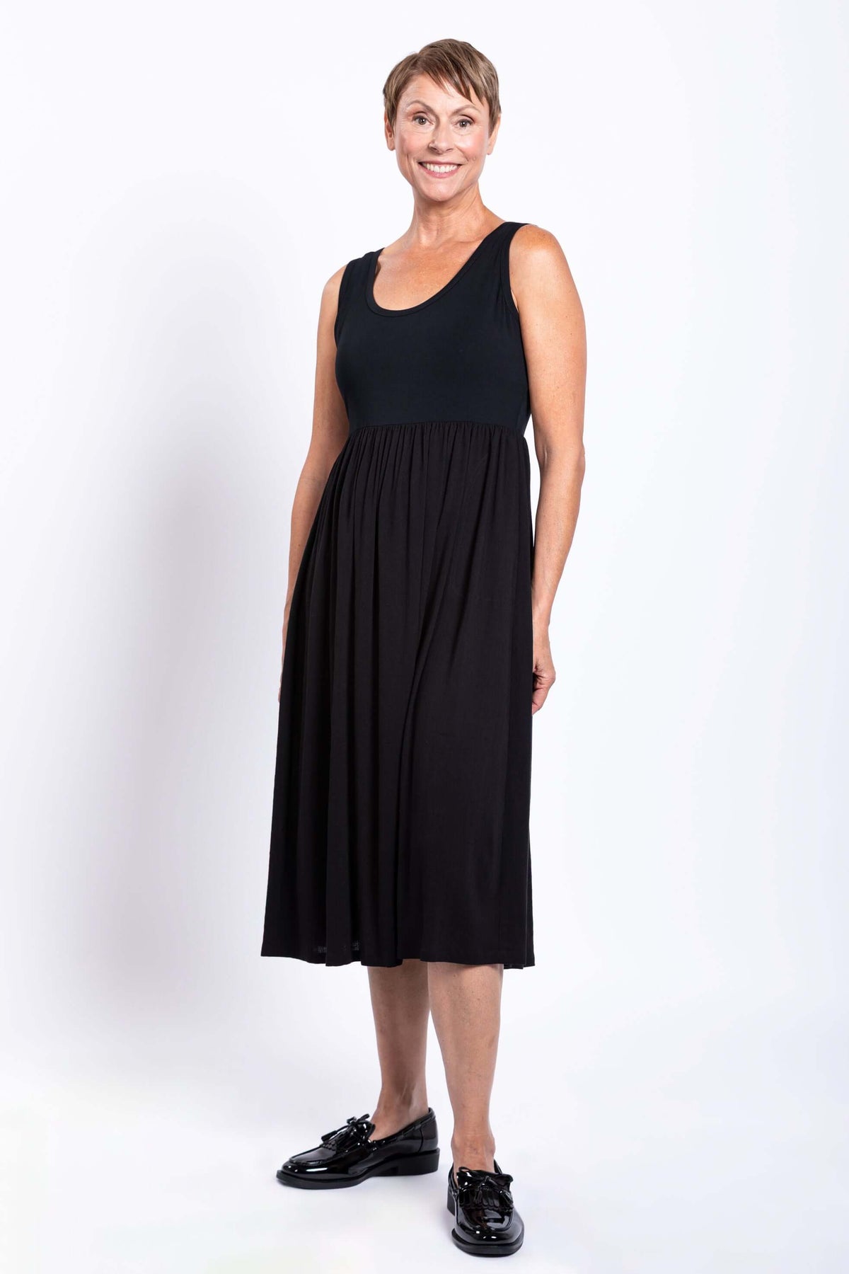 Smiling model in a black empire waist midi dress with lace-up shoes, angled view
