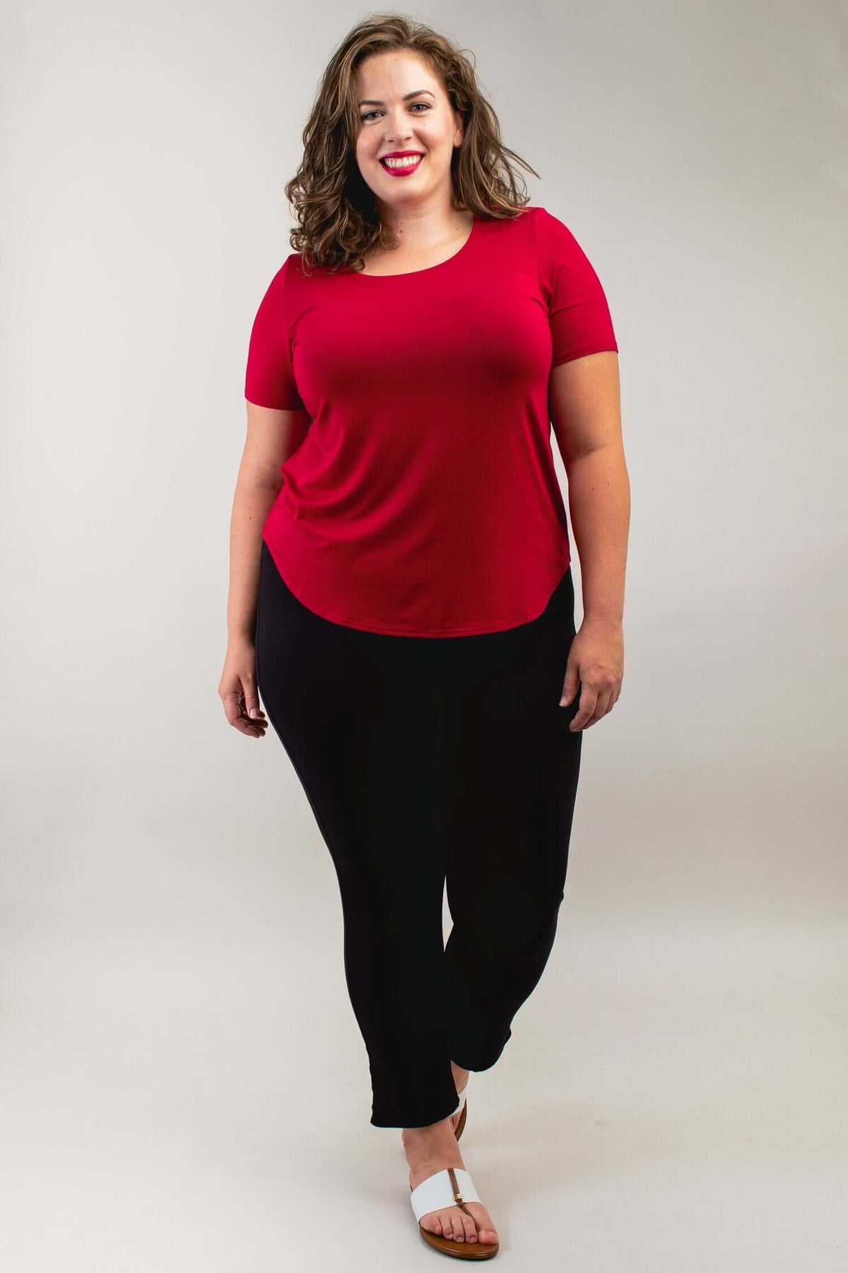 Woman standing in a crimson scoop neck tee and black pants, smiling, front view