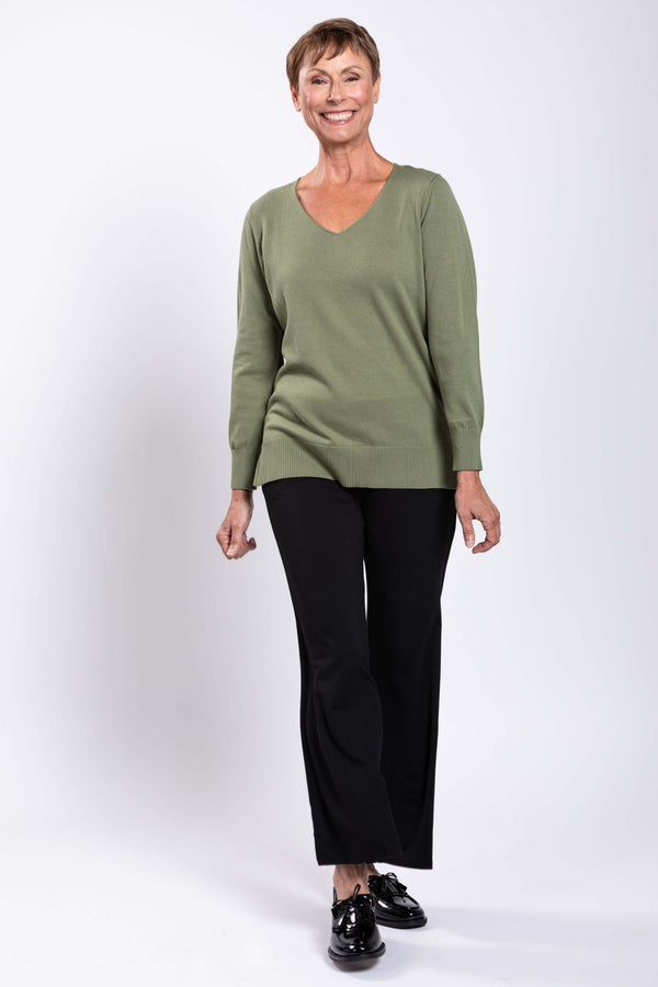 Woman smiling and wearing a sage green V-neck sweater paired with black pants and black shoes