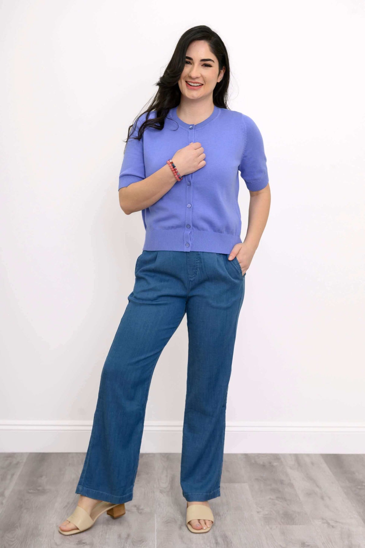Woman smiling in a short sleeve purple cardigan, hand to chest, with blue wide-leg pants