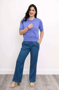 Woman smiling in a short sleeve purple cardigan, hand to chest, with blue wide-leg pants