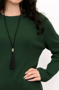 Close-up of an emerald green crew neck knit sweater styled with a long black tassel necklace, front-facing view.