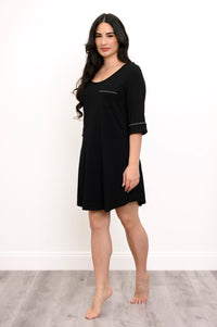 Woman standing slightly angled modeling a black sleep dress with white trim and a pocket