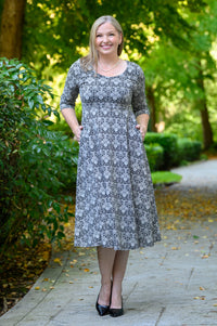 Front view of female model standing on a paved path in a wooded outdoor setting, wearing a knee-length gray midi dress with a black floral pattern, elbow-length sleeves, and black heels.