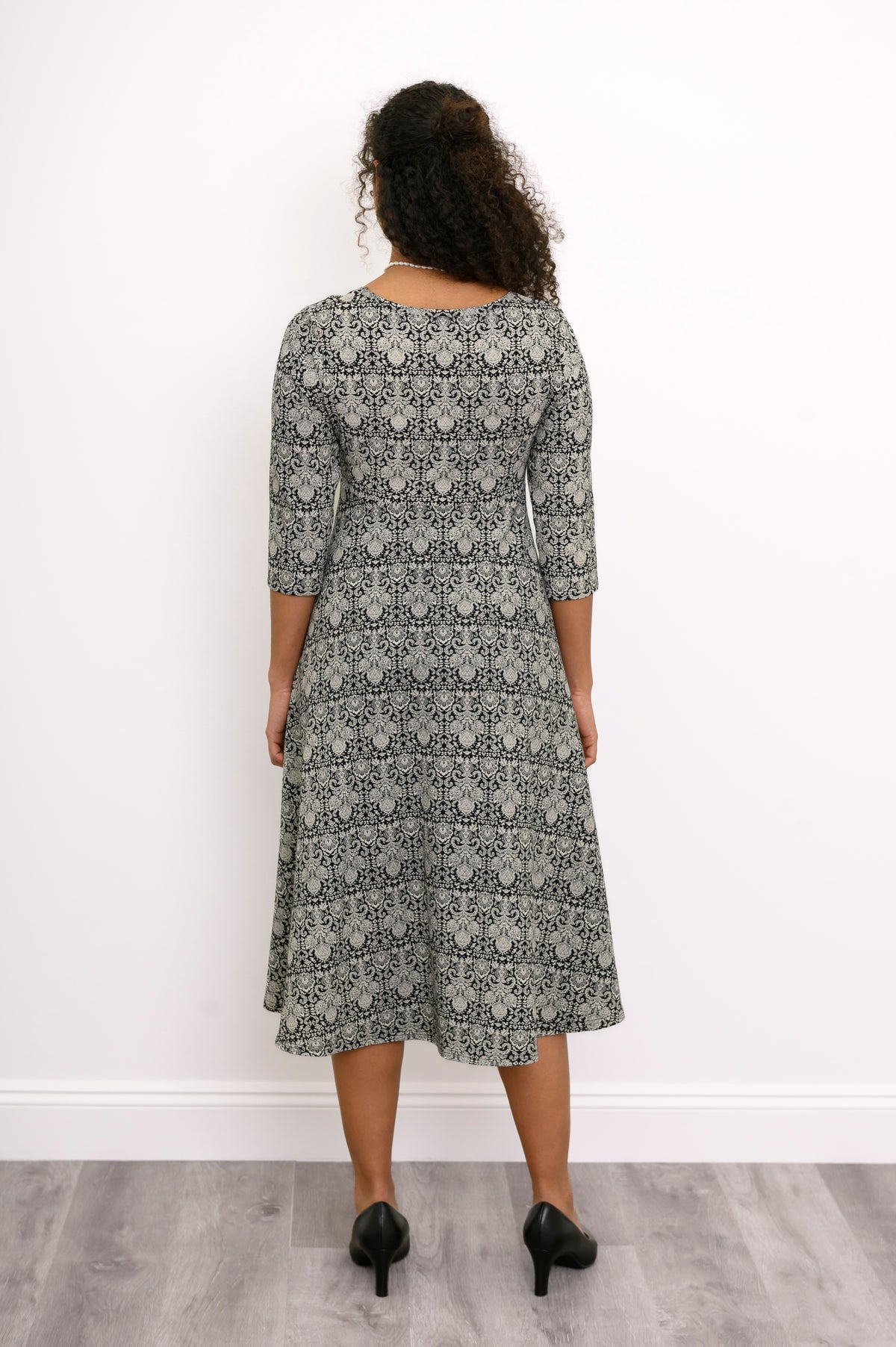 Back view of a female model wearing a grey patterned midi dress and black heels, against white wall and gray floor