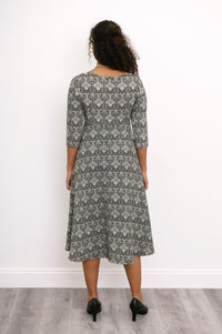 Back view of a female model wearing a grey patterned midi dress and black heels, against white wall and gray floor