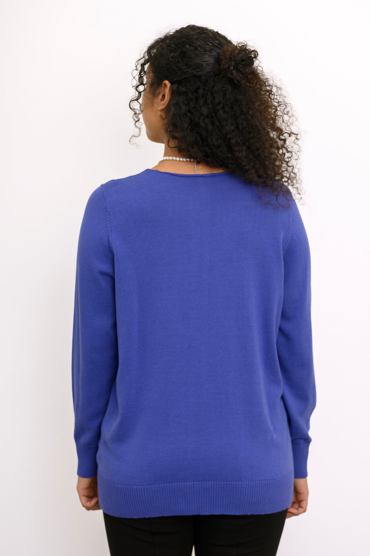 Back view of a woman wearing a hip length violet blue cotton knit sweater highlighting the clean shoulders and hem.