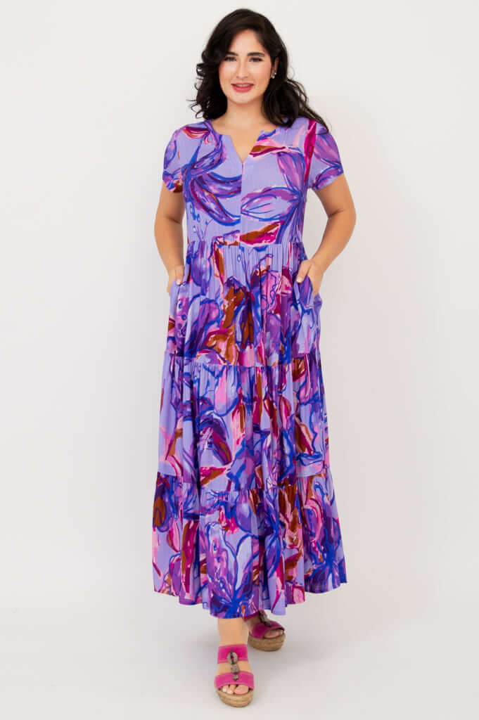 Woman wearing a vibrant purple floral maxi dress with short sleeves and tiered skirt, hands in pockets