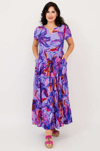 Woman wearing a vibrant purple floral maxi dress with short sleeves and tiered skirt, hands in pockets