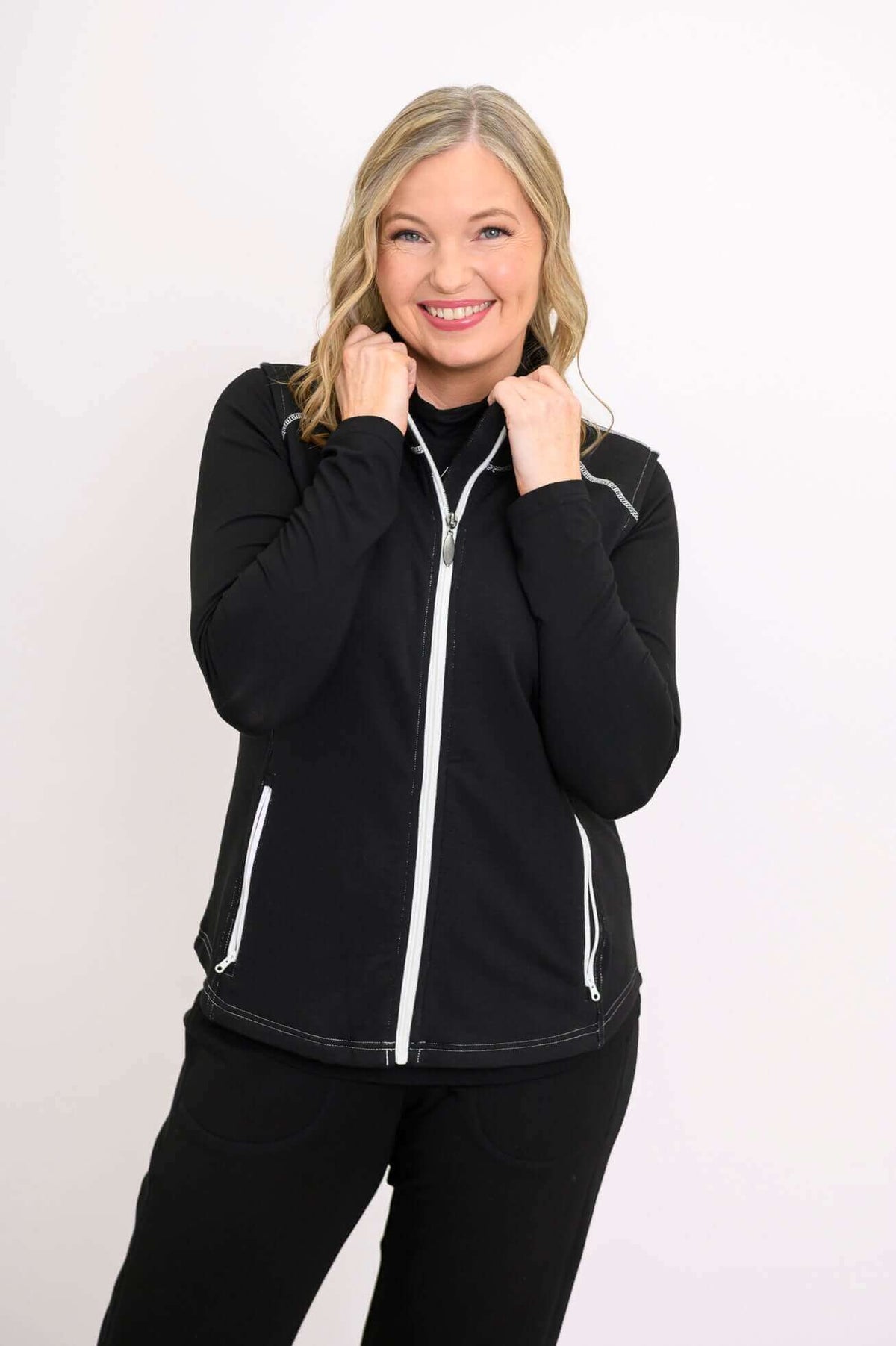 Woman smiling, holding collar of black Hartford Utility Vest with visible white zippers