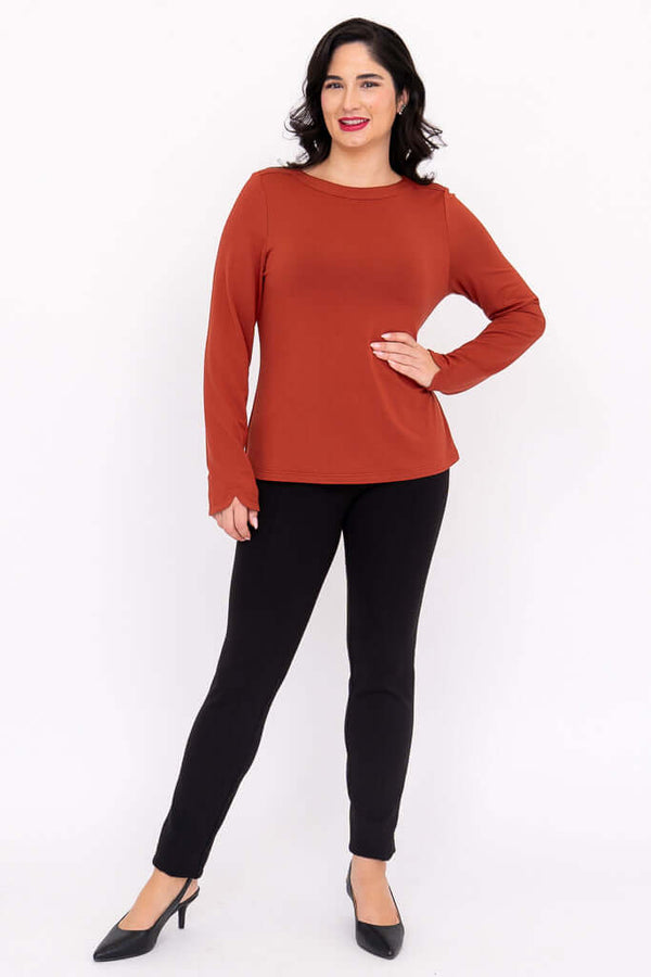 Woman in rust boat neck long sleeve top and black pants, hand on hip