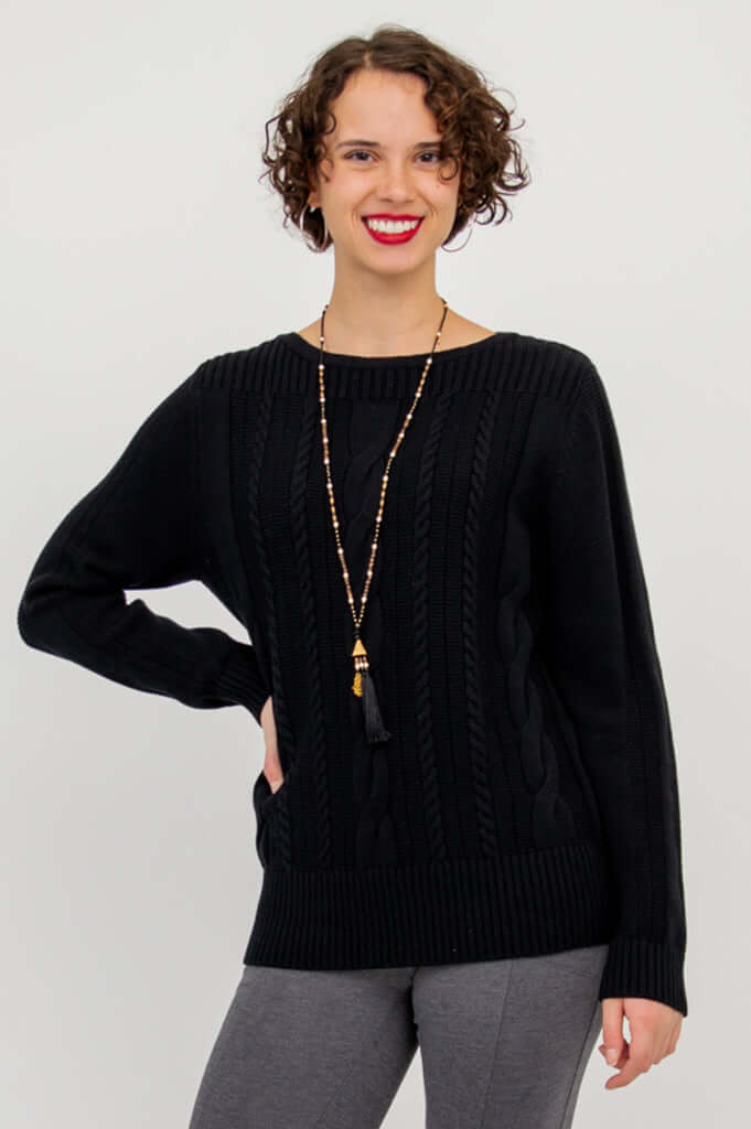 Woman wearing a black cable knit sweater with boatneck and ribbed cuffs, posing with one hand on hip
