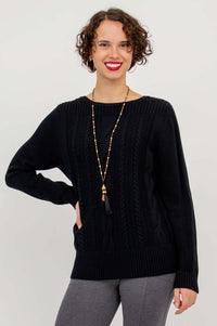 Woman wearing a black cable knit sweater with boatneck and ribbed cuffs, posing with one hand on hip