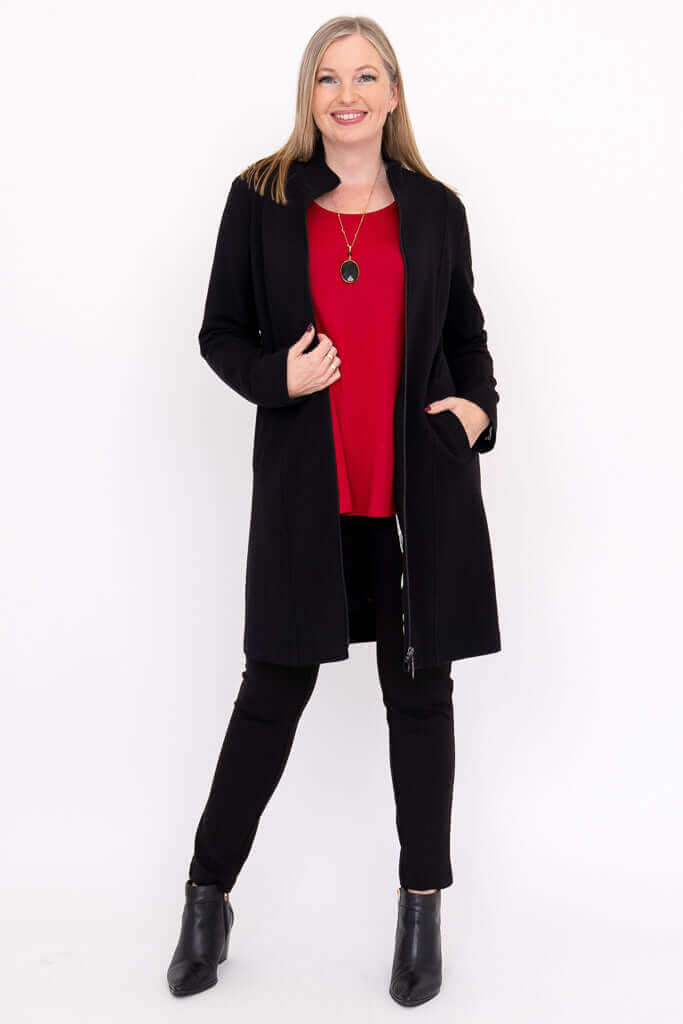 Woman in black coat over red top and black pants, holding lapel open, smiling