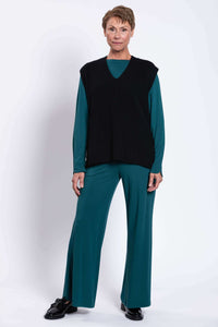 Woman standing in black sweater vest, teal pants, and teal long sleeve top