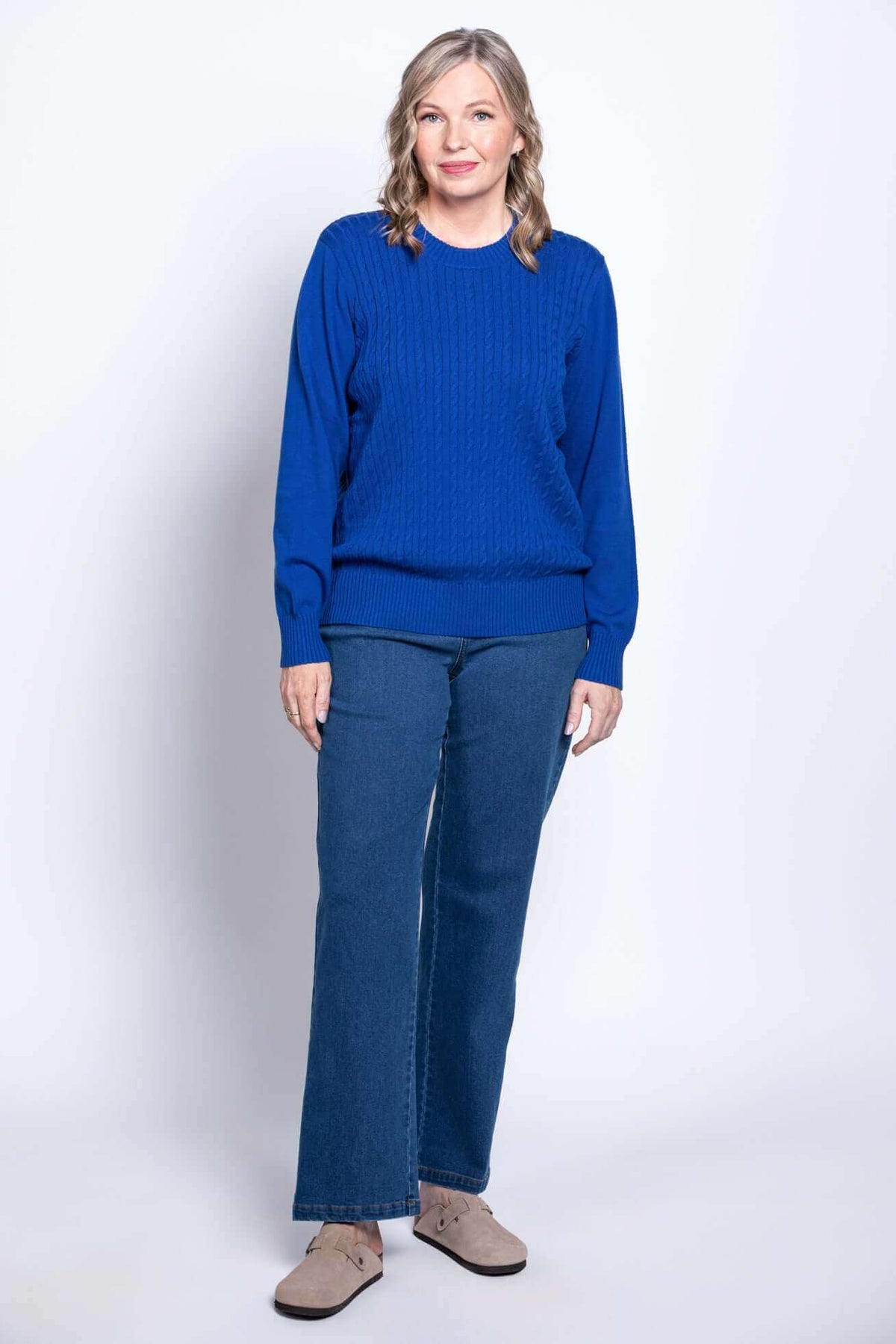 Woman in blue cable knit sweater with jeans, angled view, standing in studio setting